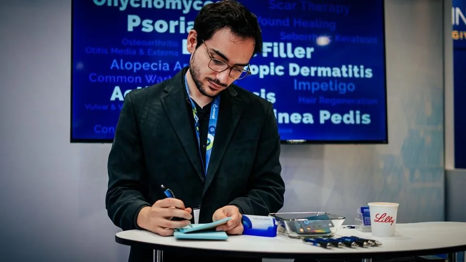 A Symbio staff member taking notes at the company booth during the AAD 2023 conference, with a sign displaying dermatology research terms like Psoriasis and Alopecia in the background.
