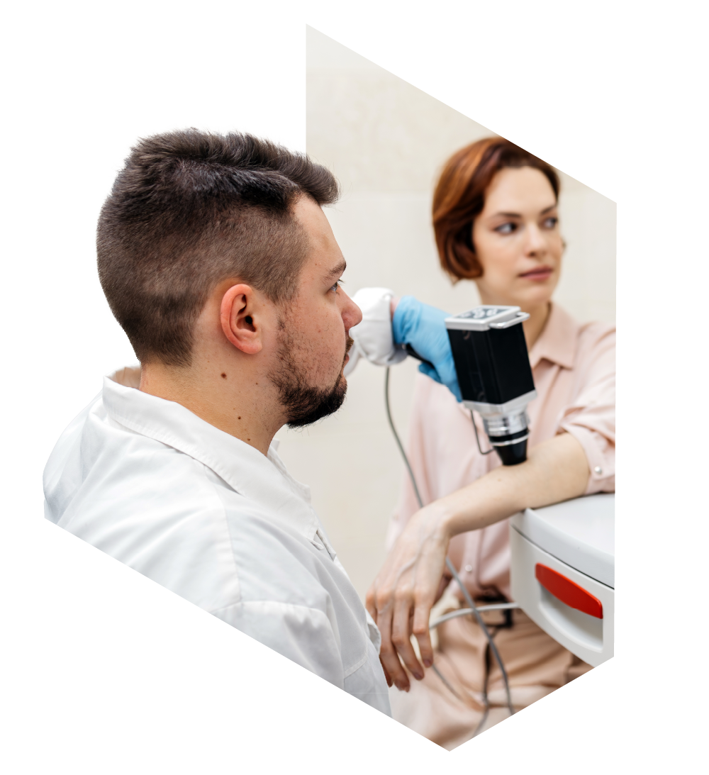 Male patient seated in a medical clinic while a female healthcare professional wearing gloves holds a handheld diagnostic device against the side of his head near the ear, appearing to perform a head or scalp examination.