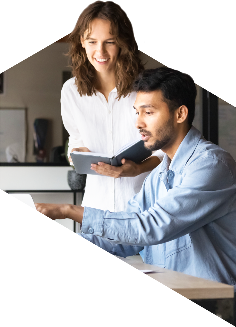 Woman standing and smiling while holding a notebook, looking at a seated man working on a laptop at a desk in an office setting, suggesting collaboration or teamwork